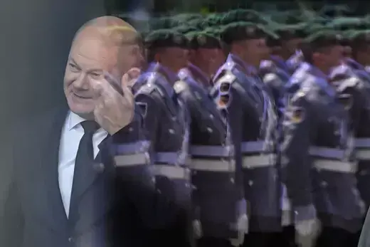German Chancellor Olaf Scholz stands behind a window in Berlin featuring a reflection of a military honor guard.