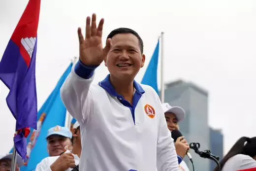 Cambodian politician Hun Manet waves to the crowd while wearing a white, long-sleeve shirt while other supporters stand in the background waving flags.
