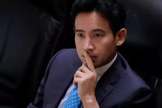 Thai Move Forward politician wears a blue blazer while sitting in a chair with his hand raised to his face.