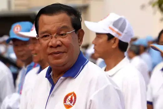 Cambodian leader wears a white polo shirt and glasses, standing in front of other party members wearing baseball hats during a political rally.