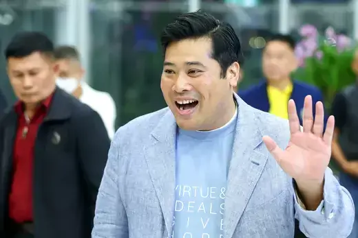 Vacharaesorn Vivacharawongse, Thailand's King Maha Vajiralongkorn's son, waves his hand while wearing a light blue t-shirt and light blue linen suit jacket. A man in a black jacket stands in the background.