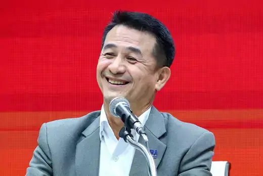 Pheu Thai leader wears a gray suit jacket and white button down shirt while sitting in front of a red background.