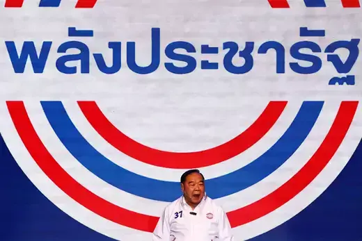 Thai politician wears a white shirt and stands in front of a backdrop with red, white, and blue lines on it.