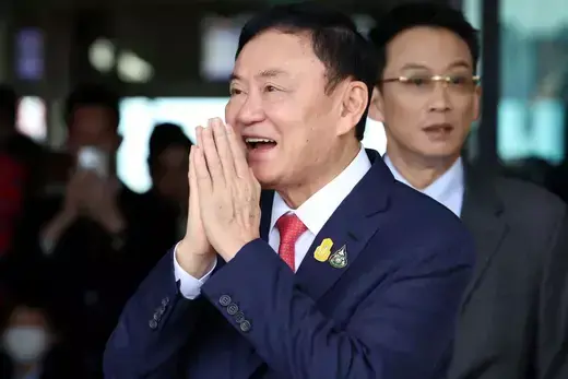 Former Thai prime minister wears a navy suit and red tie while bowing his hands in front of his face.