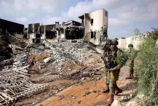 Multiple soldiers wearing green fatigues stand next to the ruins of a bombed building.