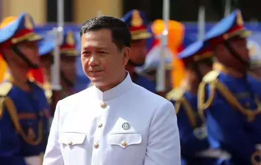 Cambodian Prime Minister Hun Manet wears a white military uniform while soldiers in blue uniforms stand at attention behind him.
