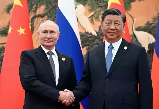 Russian president poses shaking hands with Chinese president in front of Russian and Chinese flags.