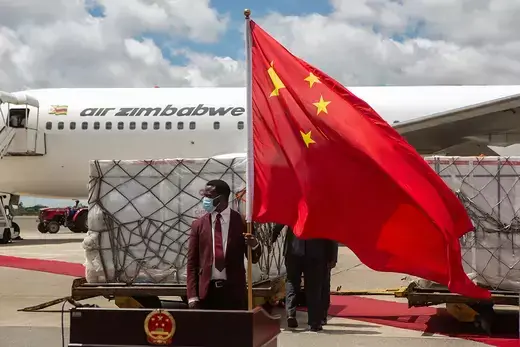 A man wearing a maroon suit hold a large Chinese flag while standing in front of a white airplane on a tarmac.