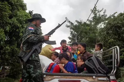 Children sit in the back of a truck while a man in army fatigues holds a gun.