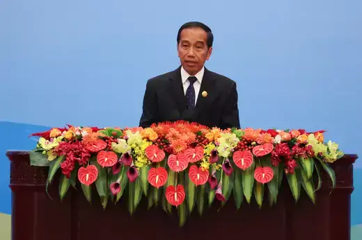 Indonesian President stands at a wooden dais overflowing with red, green, and yellow flowers behind a blue backdrop.
