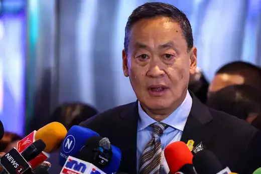 Thai Prime Minister wears a blue suit and striped tie while he speaks to the media, who hold microphones up to his face.