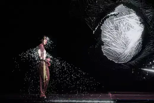 A woman stands on stage, appearing to fade away as she faces a metaphorical representation of AI. 