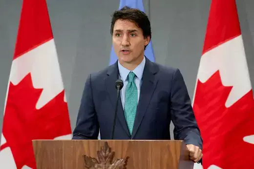 Canadian Prime Minister Justin Trudeau wears a blue suit and teal tie while standing at a wooden podium in front of two red and white Canadian flags.