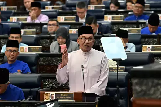 Malaysian Prime Minister wears a white shirt while giving a speech standing at his parliamentary desk. 