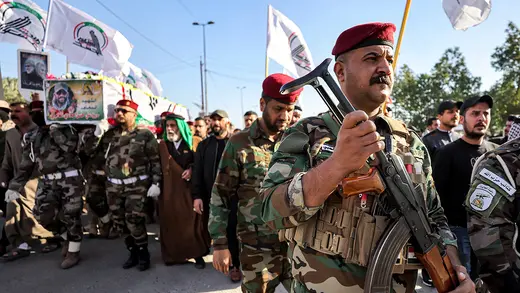A group of armed militants march in uniform during a funeral procession, carrying a coffin of a fallen troop and raising flags.