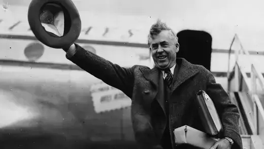 Henry Wallace standing in front of a plane waving his hat in the air as he arrives at London Airport.