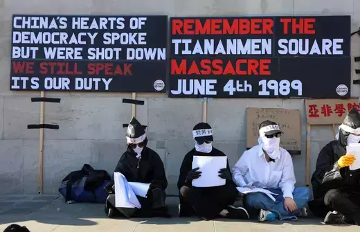 Protestors sit against the wall of a building wearing masks and sunglasses to cover their faces as black protest signs are above them