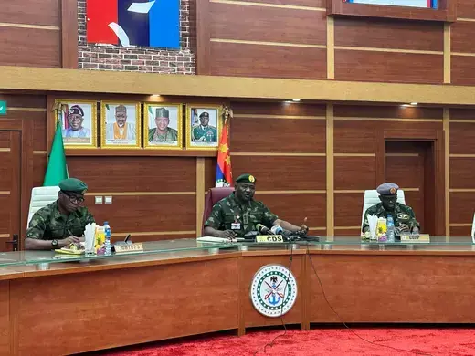 Three members of Nigeria's defense are seated next to one another in a room that has primarily wooden walls and a wooden desk.