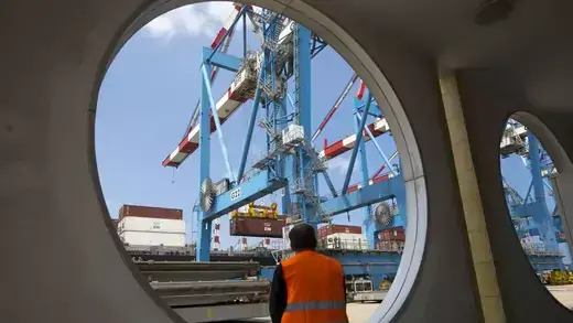 A worker sits as a crane unloads containers from a ship