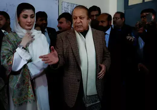 The former prime minister of Pakistan gestures with his hand while he stands with a group of people wearing a checkered blazer and white scarf.