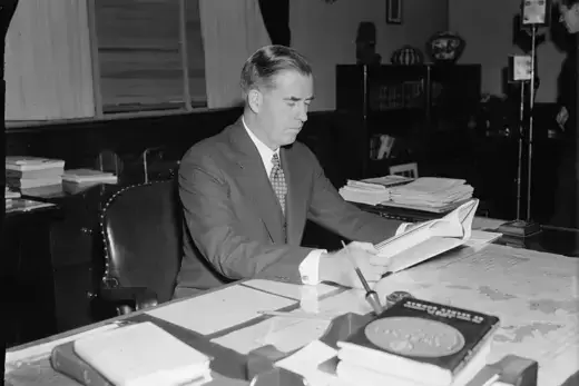 Henry Wallace as viewed sitting at a desk holding a stack of papers. 