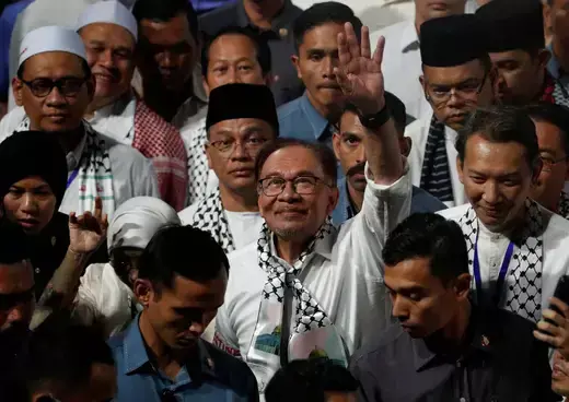 Malaysian prime mnister stands amid supporters wearing a white shirt and Palestinian keffiyeh while waving to audience.