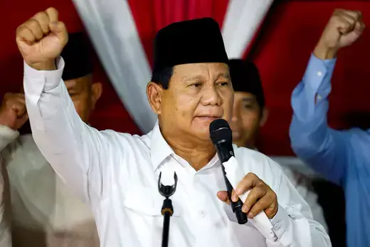 Indonesian president-elect wears a white button-down shirt and traditional black hat as he raises his fist in the air and holds a microphone in the other hand.