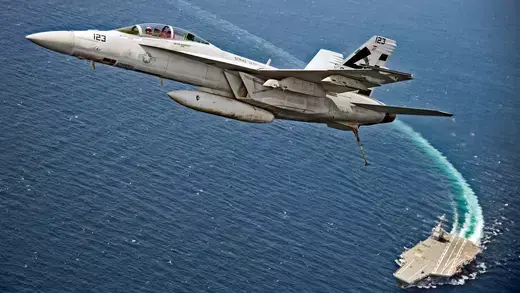 An F/A-18F Super Hornet jet flies over the USS Gerald R. Ford as the U.S. Navy aircraft carrier tests its new launch and flight arrest systems.