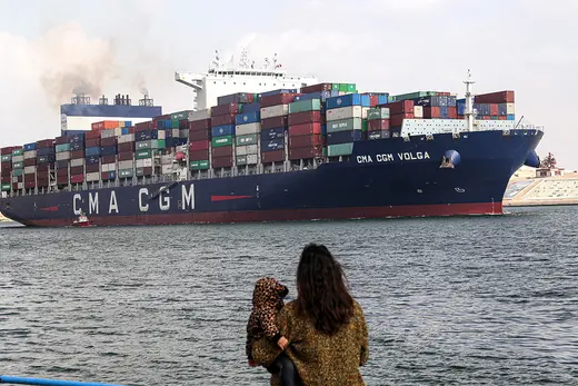 A woman holding a baby stares looks out at the Suez Canal as a cargo ship crosses.