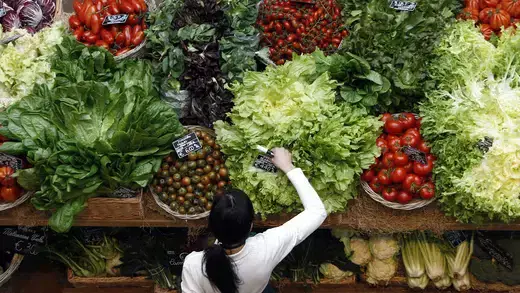 A female employee wearing a white shirt arranges pricetags at a vegetables work bench full of green leafy vegetables and red tomatoes. 