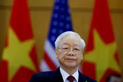 Vietnam's Communist Party General Secretary Nguyen Phu Trong stands in front of American and Viertnamese flags while looking at the camera wearing a black suit and maroon tie.