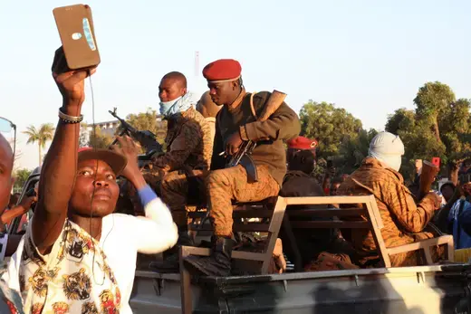 A man uses his mobile phone beside army soldiers who sit on top of a military vehicle.
