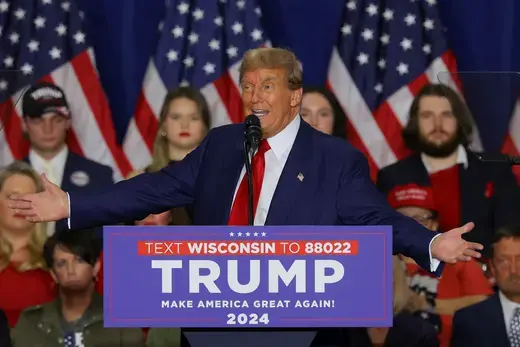 Former President Trump gestures while speaking in front of a crowd wearing a red tie and blue suit.
