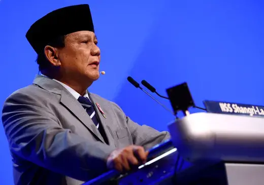 Indonesian President speaks at a podium wearing a gray suit and traditional Indonesian hat in front of a blue background.