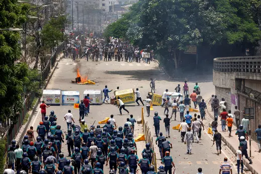 Police officers wearing riot gear face off in the street against protesters.