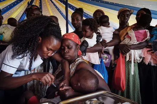 Two women vaccinate an infant in a tent surrounded by other women waiting with their children to give them the malaria shot.