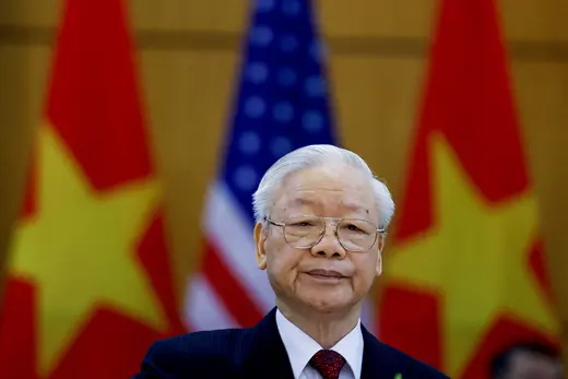 Vietnam's leader stares at the camera wearing wire rimmed glasses and a suit while standing in front of the red and yellow Vietnamese flag.