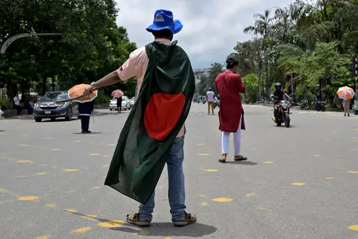 A young person stands in the middle of a street directing cars as a Bangladesh flag is draped over his shoulders.