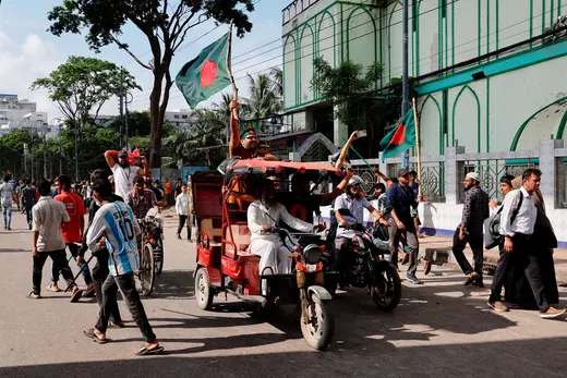 Two men drive a red rickshaw with two Bangladeshi flags in a street. 