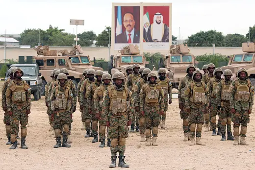 Soldiers stand in rows on a gravel lot, with large portraits of leaders behind them.