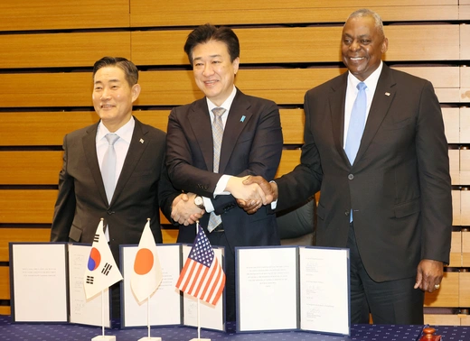 Defense ministers of Korea, Japan, and the United States wear black suits while shaking hands in front of a table with documents and small flags.