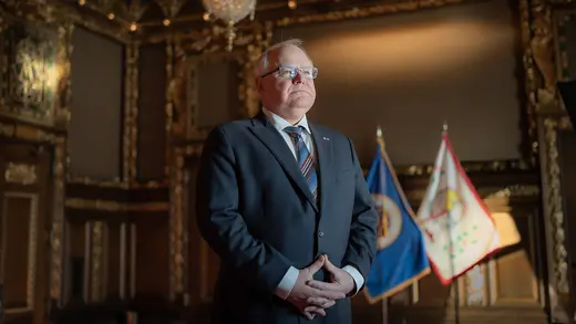 Minnesota Governor Tim Walz hold his hands to his stomach in front of the state flag.