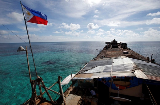 A red, white, gold, and blue Filippino flag is flown over a rusting vessel in bright blue water.