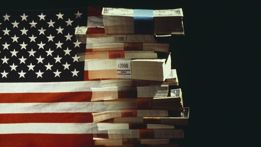 The American flag superimposed over a pile of stacked American dollar banknotes.