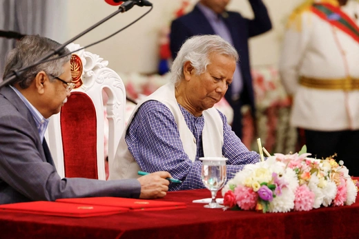 Bangladesh's interim leader sits at a table with a red velvet tablecloth and white and pink flowers while signing a document.