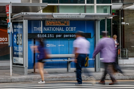 People walk in front of a bus stop with a sign saying the national debt is $104,151 per person.