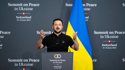 Ukrainian President Volodymyr Zelenskyy stands behind a podium with his arms raised mid-speech, a Ukrainian flag stands behind him. 