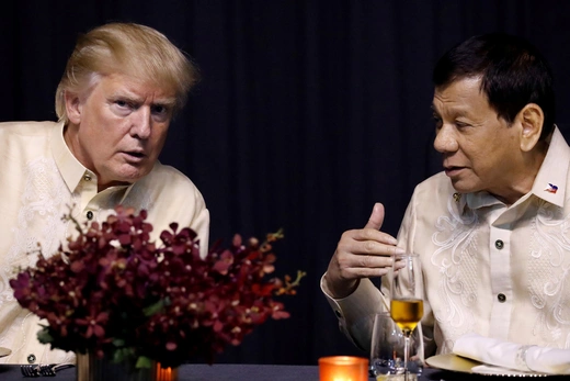 Then-President Trump wears a tan shirt while sitting next to then-Philippine President Duterte, also wearing a tan shirt, with maroon flowers on the table.