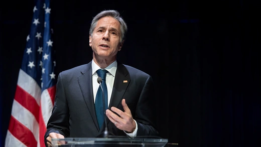 U.S. secretary of state Antony J. Blinken speaks on stage, backgrounded by the U.S. flag.