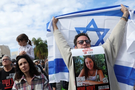 An Israeli man stands among a crowd while holding up an Israeli flag behind his head while holding a poster of an Israeli hostage by his mouth.